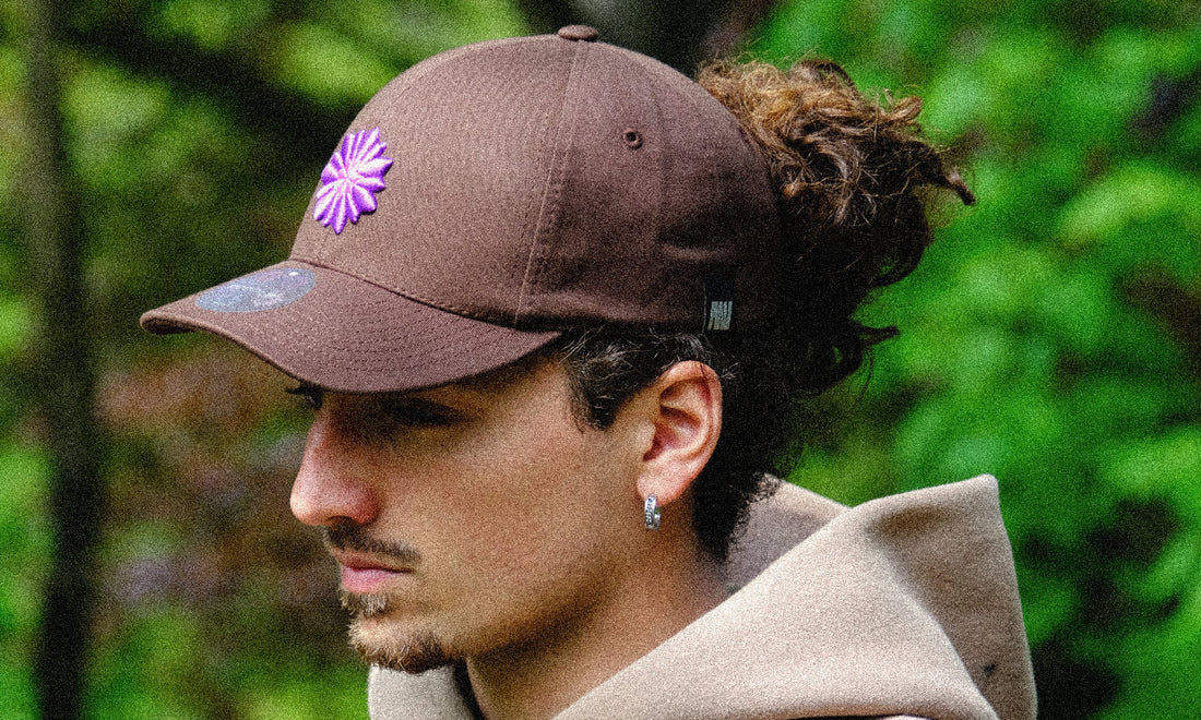 Side profile of a man wearing a brown cap with a purple Bellflower embroidery, set against a blurred green background