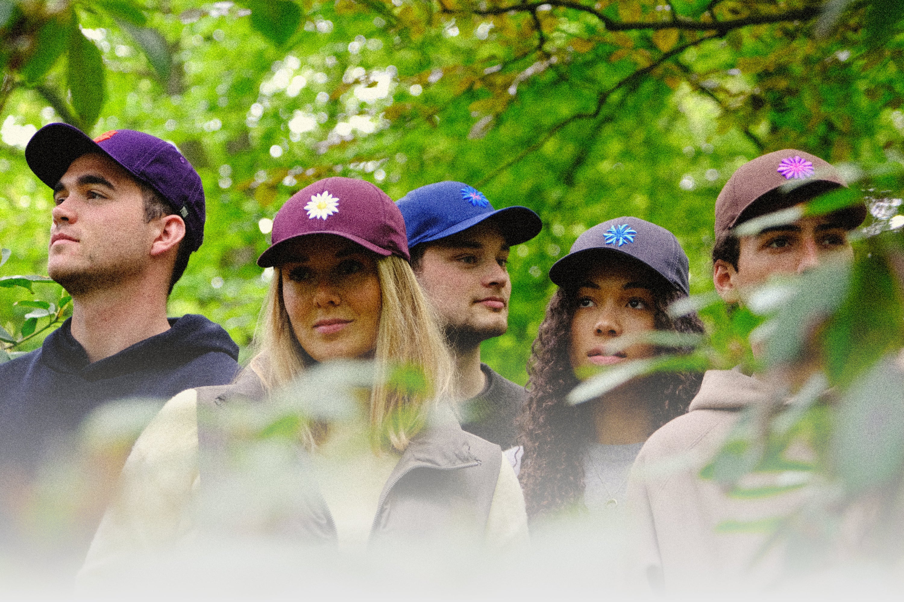 A group of four young adults wearing baseball caps with flower emblems, standing in a lush green setting. From left to right: a man in a purple cap with a red flower, a woman in a maroon cap with a white flower, a man in a blue cap with a blue and white flower, and a man in a brown cap with a pink flower. They appear thoughtful and are gazing into the distance.