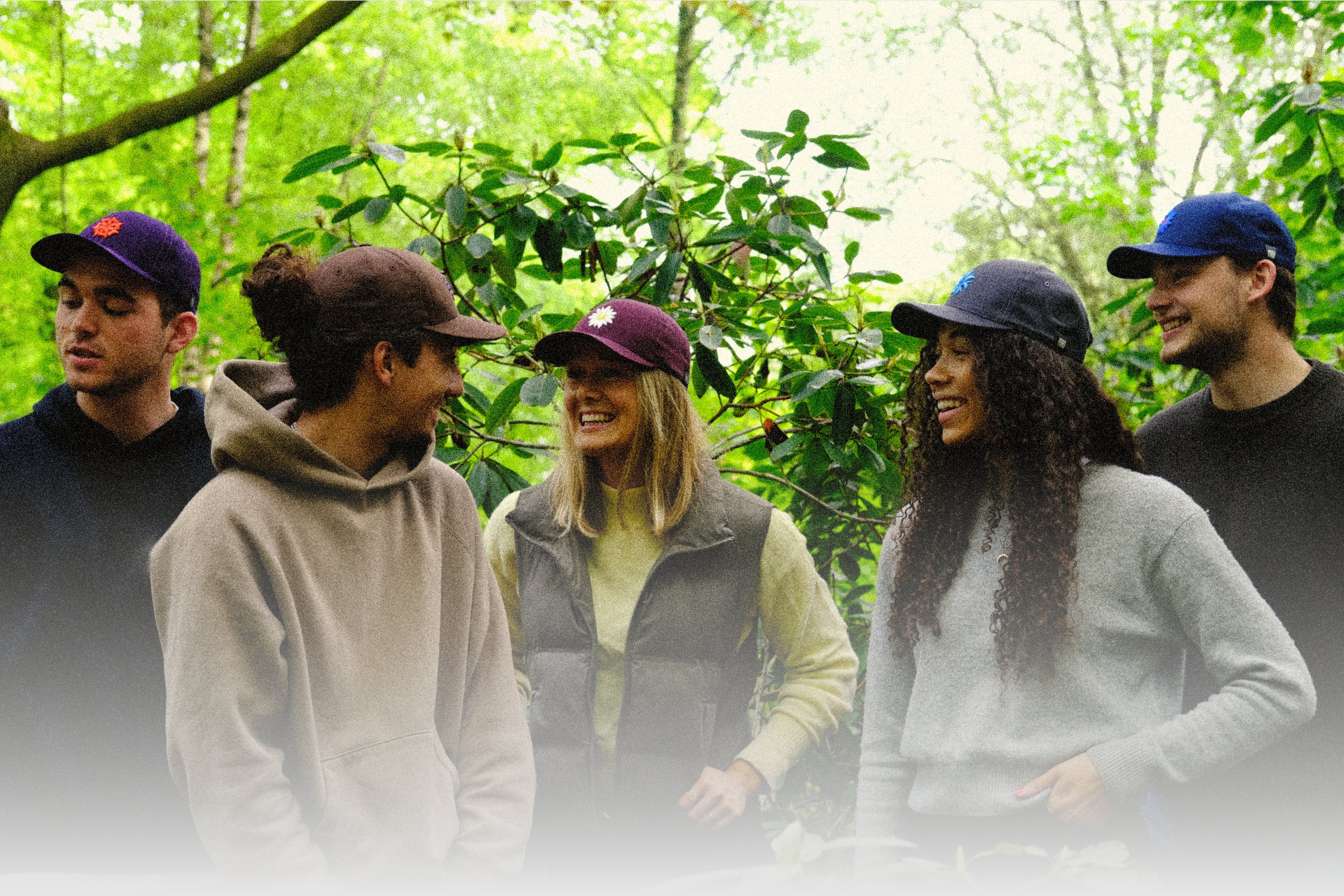 A candid moment among a group of five young adults in casual attire, laughing and conversing in a verdant outdoor setting. Each person is wearing a baseball cap with a flower emblem, in shades of purple, maroon, and blue, adding a touch of personal style to their relaxed, friendly gathering.