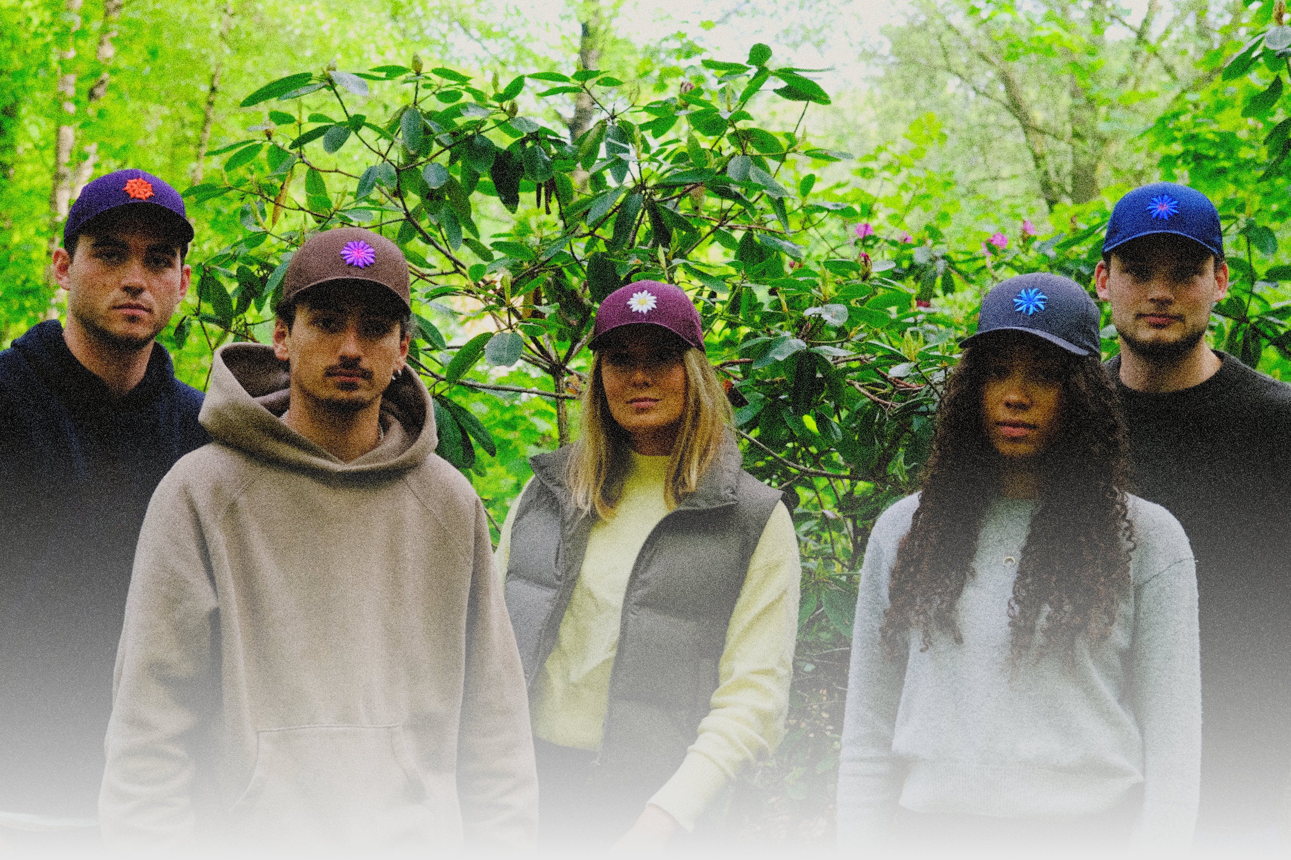 A group of four individuals standing in a line outdoors, with a lush green backdrop of leaves and foliage. Each person is wearing a distinct baseball cap with a flower emblem, showcasing colors such as purple, maroon, and blue. Their expressions are serious, and they appear to be looking directly at the viewer.